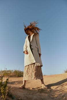 a woman in a fashionable jacket and a long skirt poses standing on the sand against the background of a clear sky. High quality photo