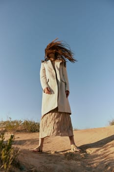 a woman in a fashionable jacket and a long skirt poses standing on the sand against the background of a clear sky. High quality photo