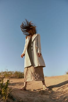 a woman in a fashionable jacket and a long skirt poses standing on the sand against the background of a clear sky. High quality photo