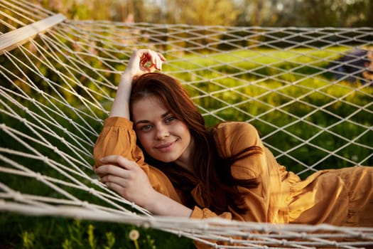 a happy woman is resting in a mesh hammock with her head resting on her hand, smiling happily at the camera with a smile, enjoying a warm day in the rays of the setting sun, lying in an orange dress. High quality photo