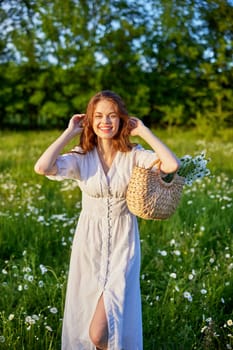 portrait of a happy red-haired woman in a light dress with a basket of flowers standing in a chamomile field and straightening her hair. High quality photo