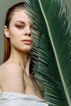 a sophisticated, elegant woman with evening makeup, stands with a green palm leaf, covering part of her face with it. Close vertical portrait, photo without retouching. High quality photo