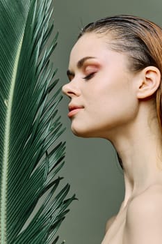 a sophisticated, elegant woman stands with a green palm leaf holding it near her face, standing sideways to the camera with her eyes closed. High quality photo