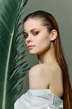 a sophisticated, elegant woman with evening makeup, stands with a green palm leaf, covering part of her face with it, dressed in a light shirt. Close vertical portrait, photo without retouching. High quality photo