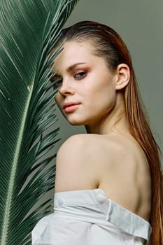 a sophisticated, elegant woman with evening makeup, stands with a green palm leaf, covering part of her face with it, dressed in a light shirt. Close vertical portrait, photo without retouching. High quality photo