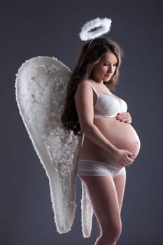Pretty pregnant woman in angel costume posing on gray background