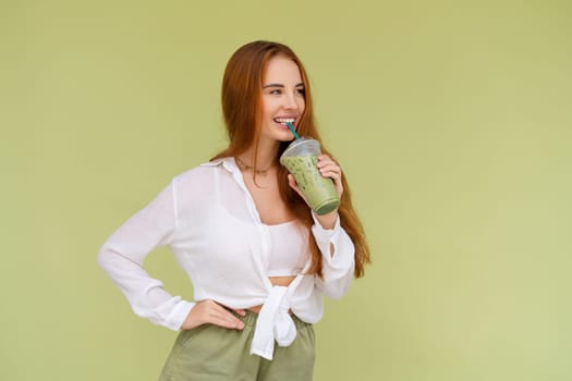 Beautiful red hair woman in casual shirt on green background positive vibes enjoying fresh ice green matcha tea with milk at hot summer day