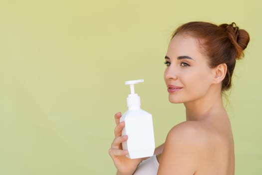 Beauty portrait of young topless red hair woman with bare shoulders on green background with perfect skin holding bottle of shampoo, body lotion