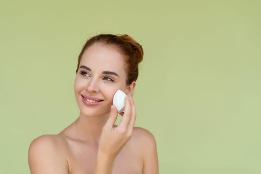 Beauty portrait of young topless red hair woman with bare shoulders on green background with makeup foundation sponge