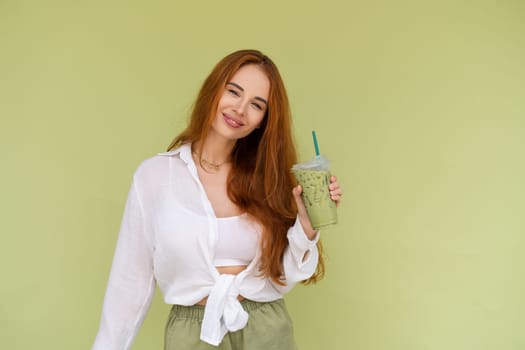 Beautiful red hair woman in casual shirt on green background positive vibes enjoying fresh ice green matcha tea with milk at hot summer day