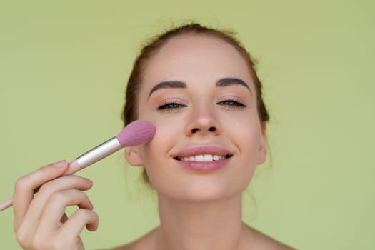 Beauty portrait of young topless red hair woman with bare shoulders on green background with perfect skin and natural makeup holds blush brush