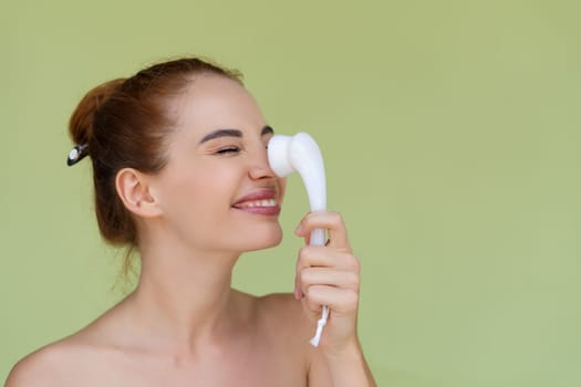 Beauty portrait of young topless red hair woman with bare shoulders on green background with perfect skin and natural makeup holds face cleansing brush