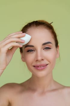 Beauty portrait of young topless red hair woman with bare shoulders on green background with makeup foundation sponge