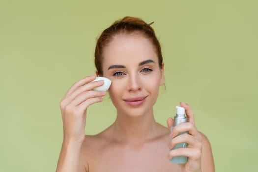 Beauty portrait of young topless red hair woman with bare shoulders on green background holds serum for youth and skin hydration and sponge