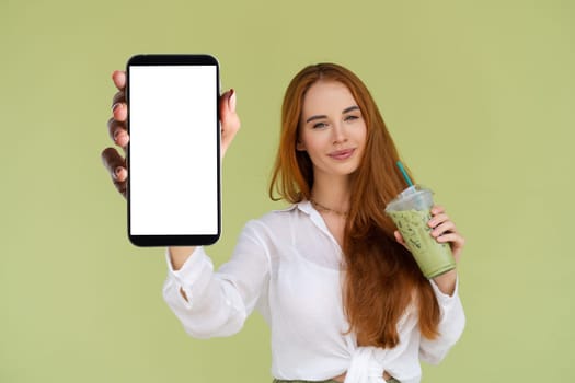 Beautiful red hair woman in casual shirt on green background holds a phone with a blank white screen and matcha ice green tea latte