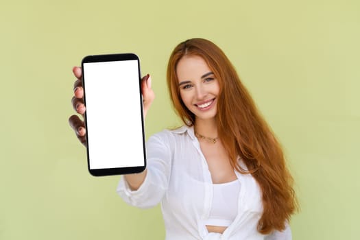 Beautiful red hair woman in casual shirt on green background holds a phone with a blank white screen