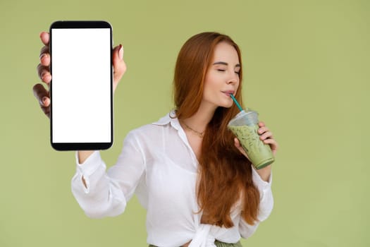 Beautiful red hair woman in casual shirt on green background holds a phone with a blank white screen and matcha ice green tea latte