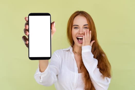 Beautiful red hair woman in casual shirt on green background holds a phone with a blank white screen