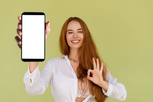 Beautiful red hair woman in casual shirt on green background holds a phone with a blank white screen