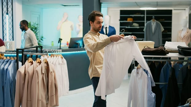 Young person buying casual wear in department store, caucasian man looking at hangers filled with modern clothes. Fashion market client checking boutique products on display, commercial activity.