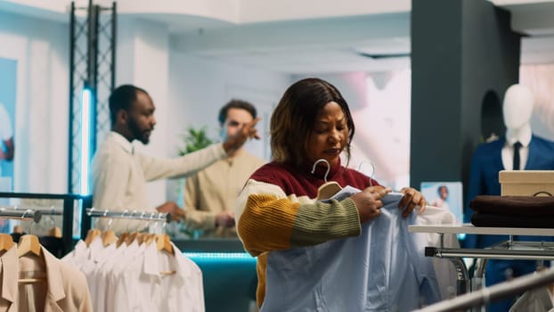 Young customer looking at clothes on hangers, analyzing merchandise in clothing fashion shop. Modern woman checking fashionable formal wear on racks, commercial activity in department store.