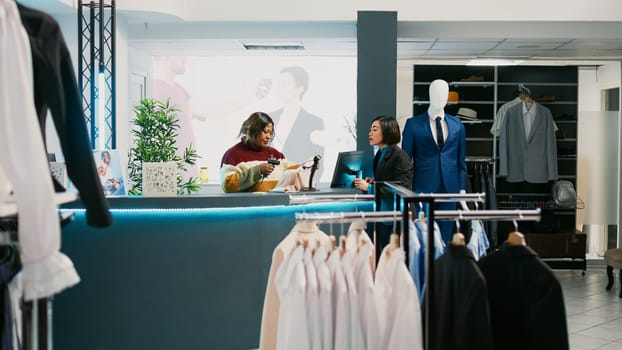 Asian person paying for merchandise at cash register, using card at pos terminal in clothing store. Young happy woman talking to retail worker, buying trendy formal wear. Commercial activity.