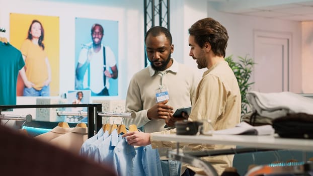 Shop employee helping customer with modern trends to buy new merchandise in clothing store. Young man talking to assistant and examining formal wear on in boutique. Handheld shot.