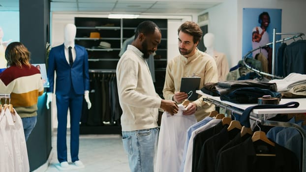 African american man checking merchandise on hangers, asking store assistant about new fashion collection. Young client talking to mall employee about clothes in retail shop boutique.