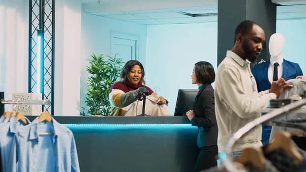 Asian person buying fashion collection in boutique shop, making electronic payment at pos terminal. Young client talking to store employee at cash register before transaction, small business.