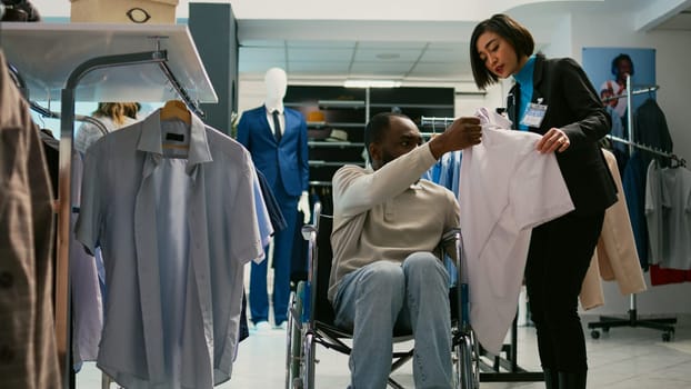 Young adult wheelchair user buying modern clothes in clothing store, suffering from physical disability at boutique. Male shopper with impairment buying new casual fashion wear, shopping mall.