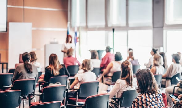 Business and entrepreneurship symposium. Female speaker giving a talk at business meeting. Audience in conference hall. Rear view of unrecognized participant in audience.