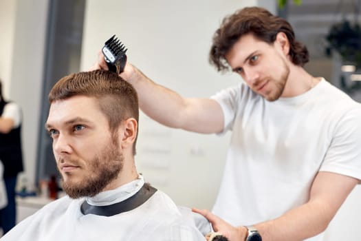 Barber trim hair with clipper on handsome bearded man in barber shop.