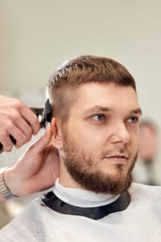 Barber trim hair with clipper on handsome bearded man in barber shop.