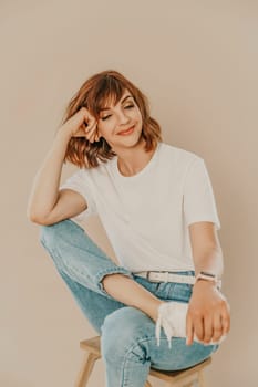 Portrait of brunette white t-shirt. A woman sits on a chair and looks at the camera, business portrait on a beige background, dressed in a white t-shirt and jeans