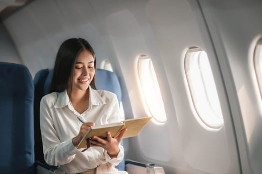 Asian young woman take notes in a notebook sitting near windows at first class on airplane during flight,Traveling and Business concept..