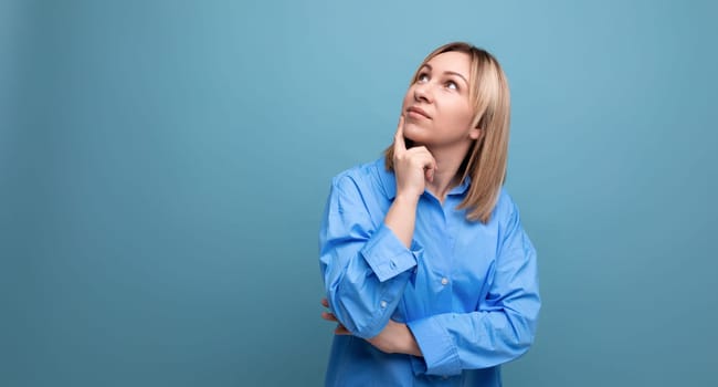 bright blond young woman in a casual shirt looks up thoughtfully on a blue background with copy space.