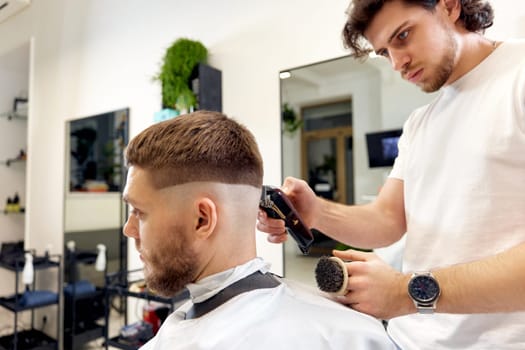 Barber trim hair with clipper on handsome bearded man in barber shop.