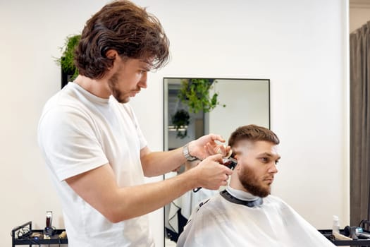 Barber trim hair with clipper on handsome bearded man in barber shop.