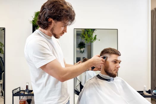 Barber trim hair with clipper on handsome bearded man in barber shop.