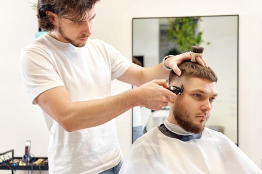 Barber trim hair with clipper on handsome bearded man in barber shop.