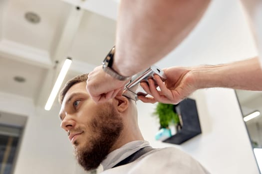 Barber trim hair with clipper on handsome bearded man in barber shop.