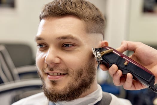 Barber trim hair with clipper on handsome bearded man in barber shop.