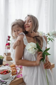 A little blonde girl with her mom on a kitchen countertop decorated with peonies. The concept of the relationship between mother and daughter. Spring atmosphere