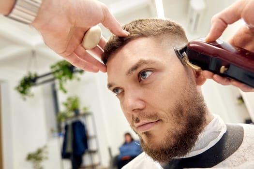 Barber trim hair with clipper on handsome bearded man in barber shop.