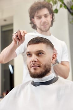 hairdresser does haircut for caucasian bearded man using comb and grooming scissors in barber shop.