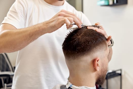 close-up, hairdresser does haircut for caucasian bearded man using comb and grooming scissors in barber shop.