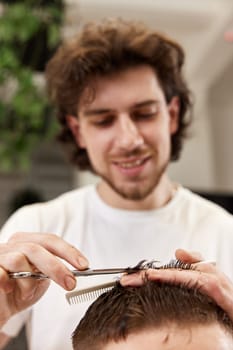 hairdresser does haircut for caucasian bearded man using comb and grooming scissors in barber shop.