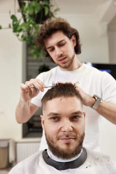 hairdresser does haircut for caucasian bearded man using comb and grooming scissors in barber shop.