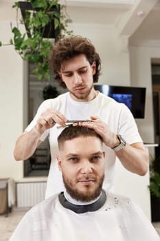 hairdresser does haircut for caucasian bearded man using comb and grooming scissors in barber shop.