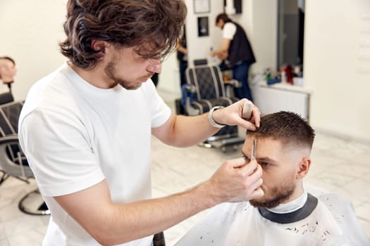 hairdresser does haircut for caucasian bearded man using comb and grooming scissors in barber shop.